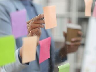 person writing on coloured post-it notes on a glass office wall while holding a takeaway coffee cup